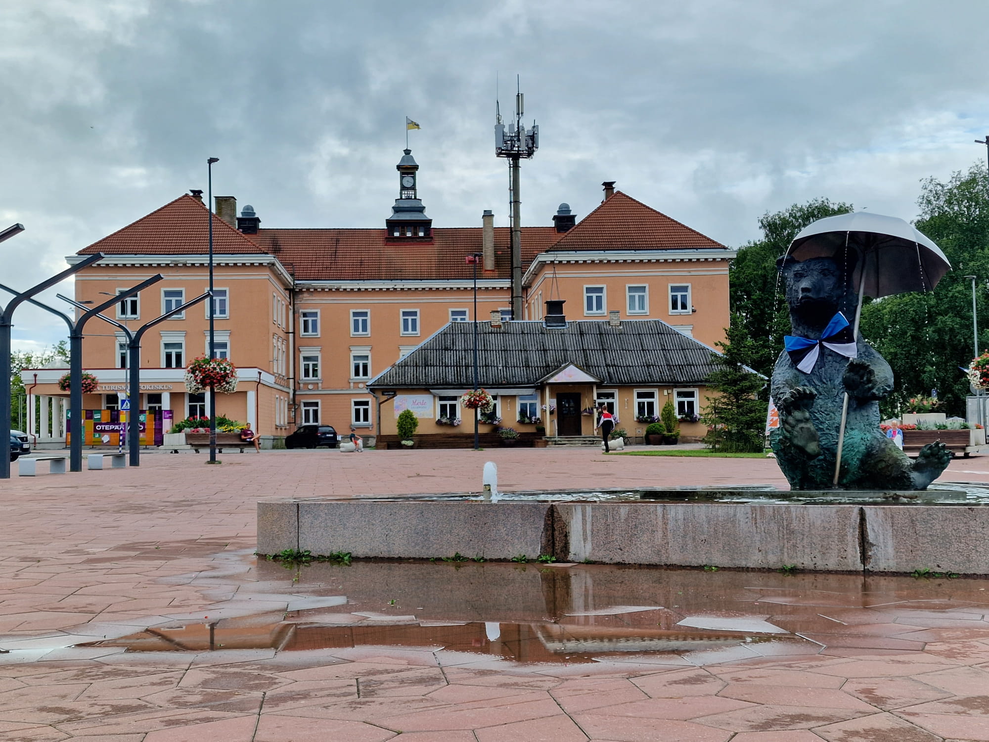 The photo shows Otepää Central Square on a rainy day. In the foreground there is a shallow fountain basin with a low granite edge; large puddles and reflections are visible on the wet paving. On the right, next to the fountain, there is a large bear sculpture wearing a bow tie and holding an umbrella.