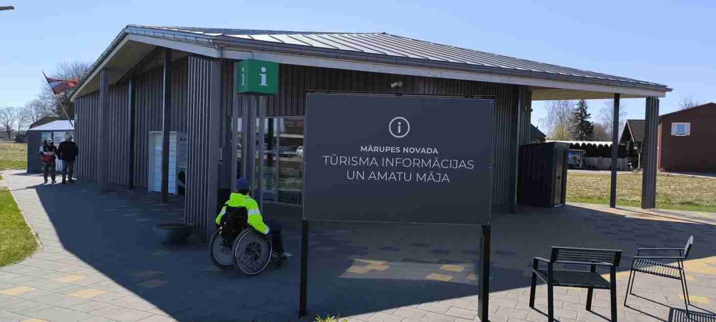 Vienstāvu ēka, Latvijas karogs pie ēkas piestiprināts, priekšā uzraksts un cilvēks ratiņkrēslā
