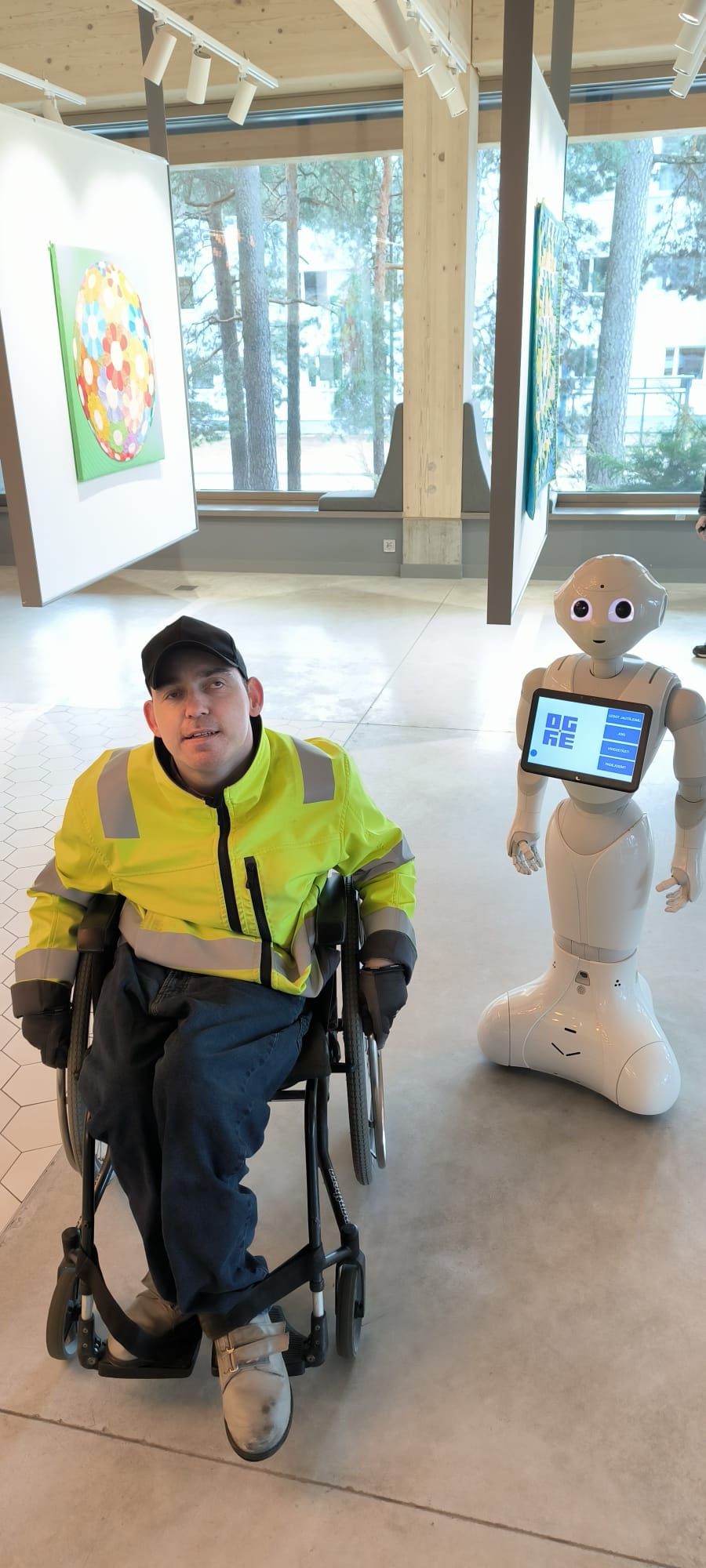 Voldemārs pozē ar baltas krāsas robotu, kuram priekšā ir displejs