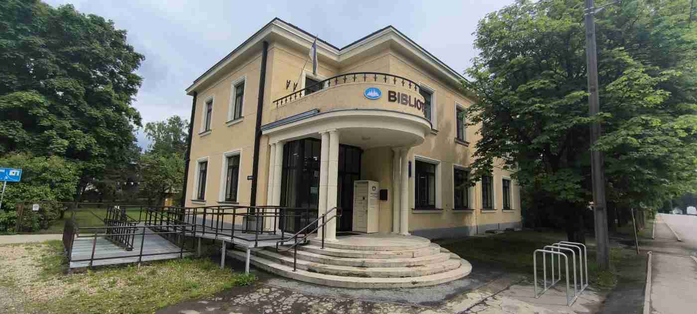 Dzeltenas krāasa divstāvu ēka ar uzrakstu Bibliotēka, kurai sānā ir uzbrauktuve, no priekšpuses kāpnes