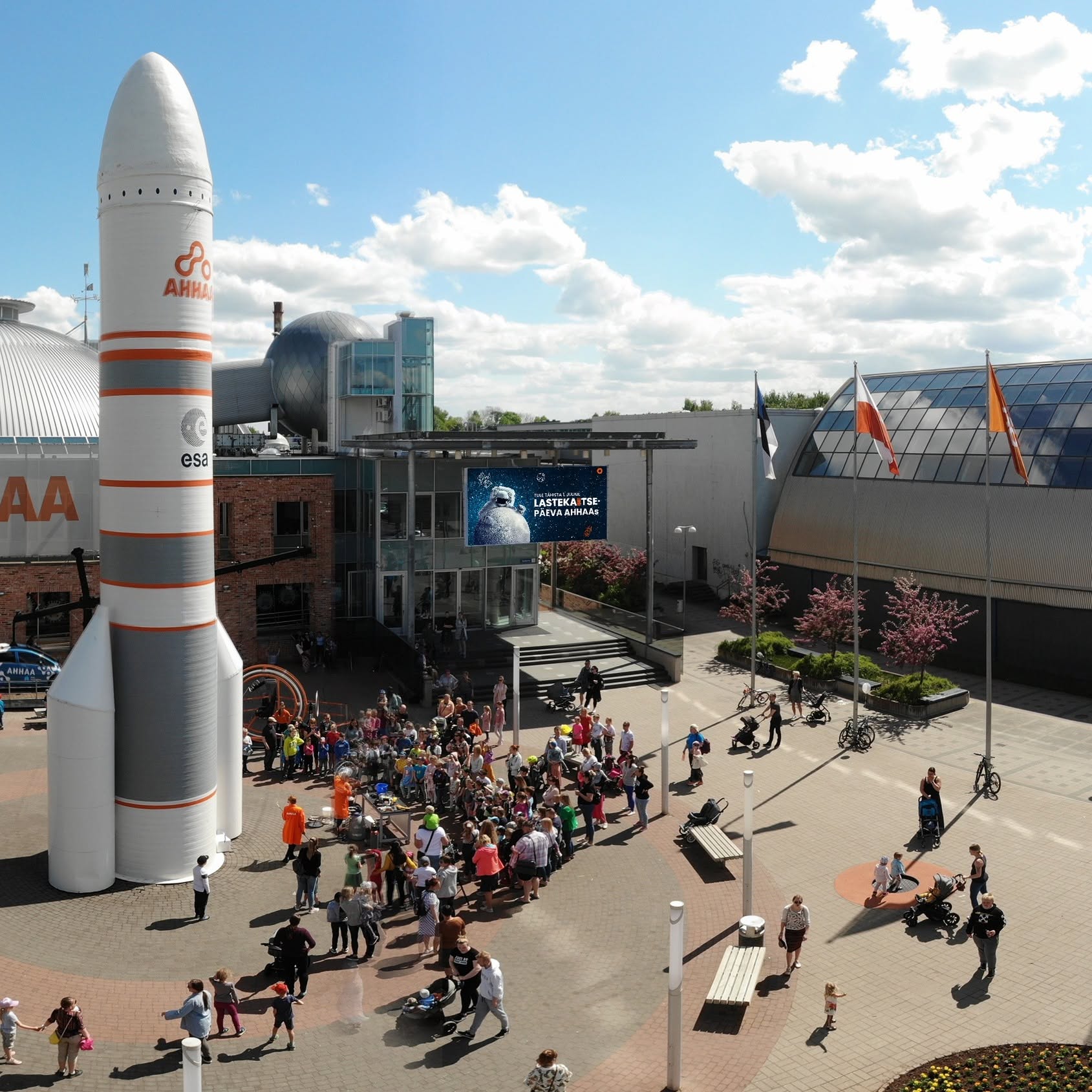 Pildil on õuest, kõrgemalt tehtud pilt Ahhaa keskuse esimest alast ja hoonest. Hoone ees on platsil palju rahvast ja Ahhaa hoone ees on ka suur rakett, mis on keskusest kõrgem.