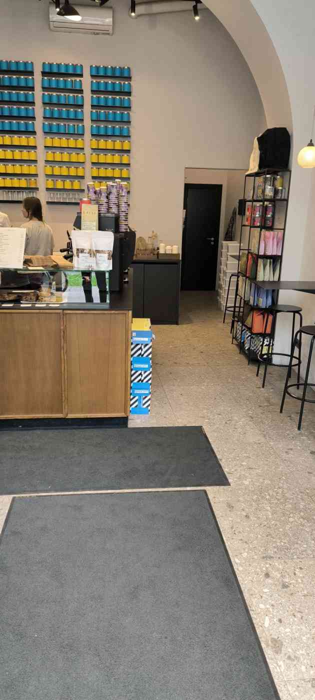 Kafejnīcas iekšpusē pie sienas daudz zilā, dzeltenā krāsā bundžas, uz grīdas paklāji, zema lete, galds, krēsli, plaukts