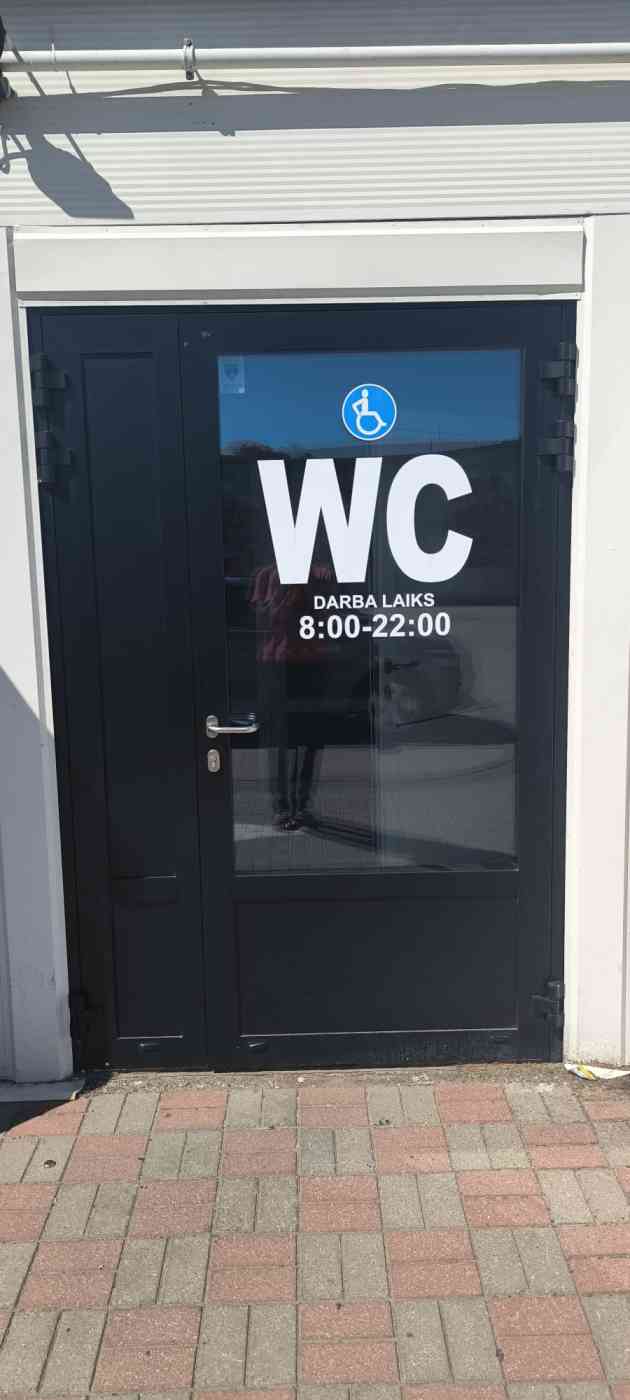 Tualetes ieejas durvis. Uz durvīm uzraksts WC un darba laiks 8:00-22:00. Virs uzraksta zilas krāsas cilvēka ratiņkrēslā zīme