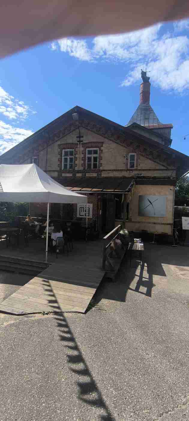 Divstāvu ēka ar terasi un uzbrauktuvi no koka. Uz terases baltas krāsas liels auduma saulessargs, galdi, krēsli