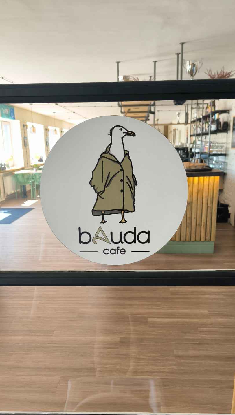Uz stiklotā durvīm attēls ar uzrakstu bAuda cafe. Fonmā redzama iekštelpa