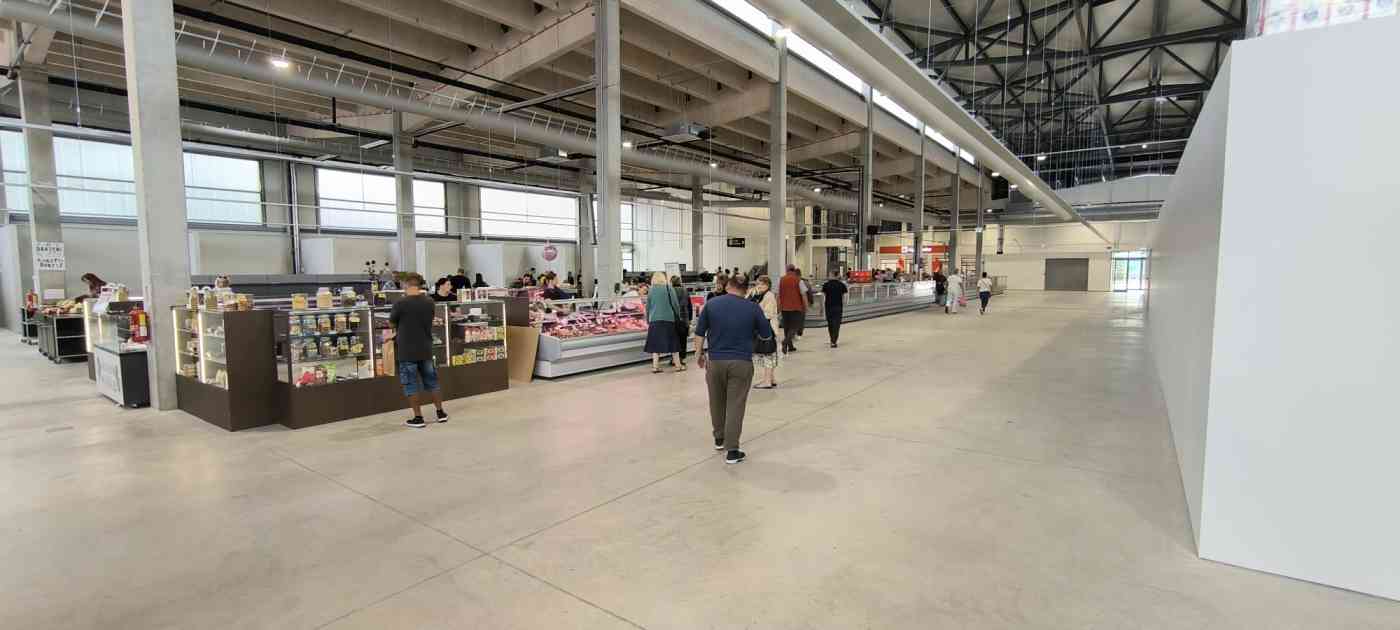 Tirgus paviljona iekštelpa ar vairākām letēm. Betona stabi, ventilācijas caurules, pelēcīgas lielas grīdas flīzes.