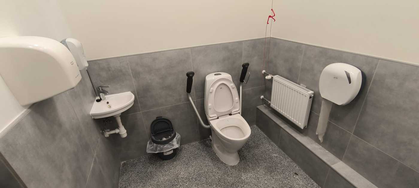 Tualetes telpā balts pods, izlietne, bērnu pārtinamais galdiņš, radiators, tualetes papīra turētājs, papīra dvieļu turētājs, šķidrās ziepes. Melnā krāsā miskaste